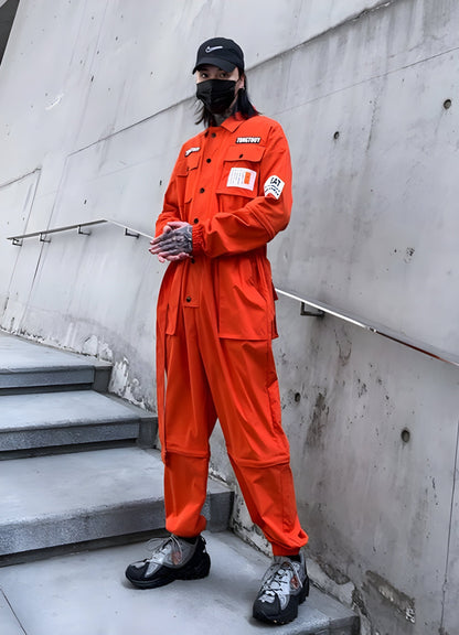 The outfit features a mix of technical fabrics and utility elements, including a high-collar jacket, cargo pants, and rugged boots, all in shades of orange.